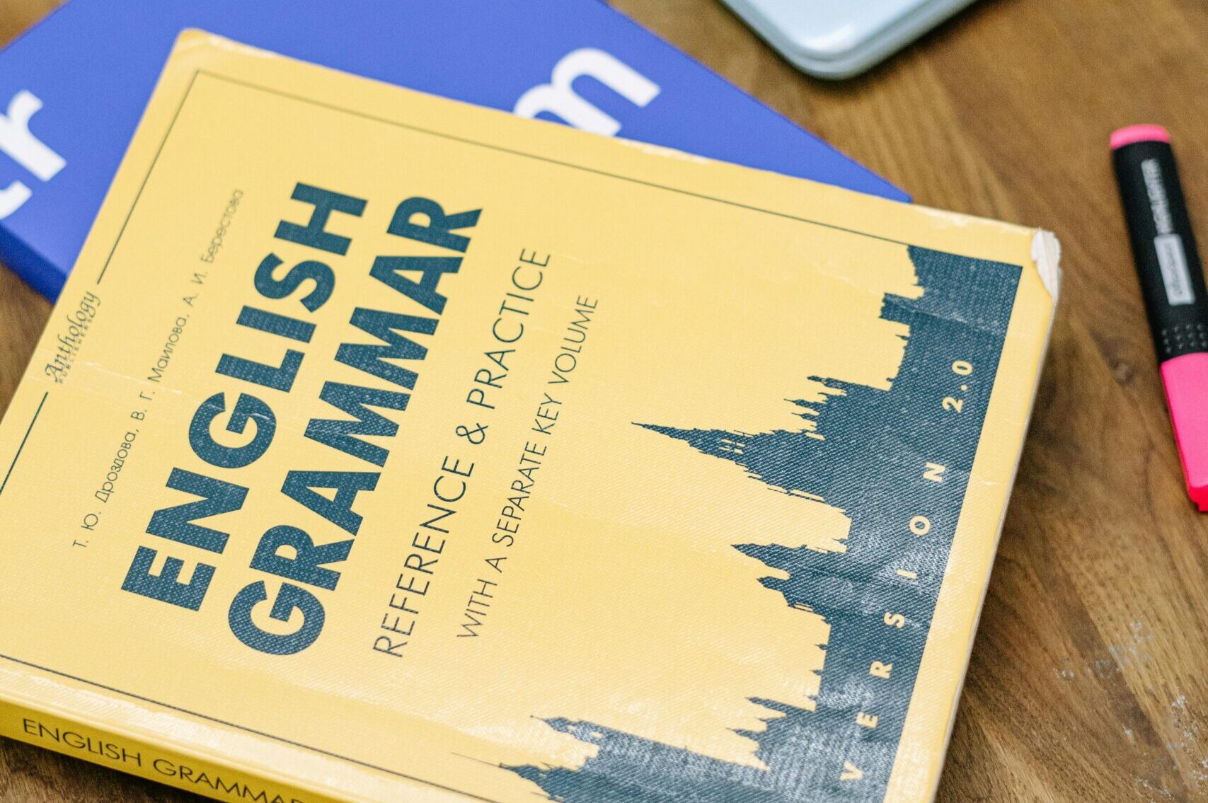 pexels-photo-5676740-5676740 High-angle view of an English grammar book and a laptop with stickers on a wooden desk.